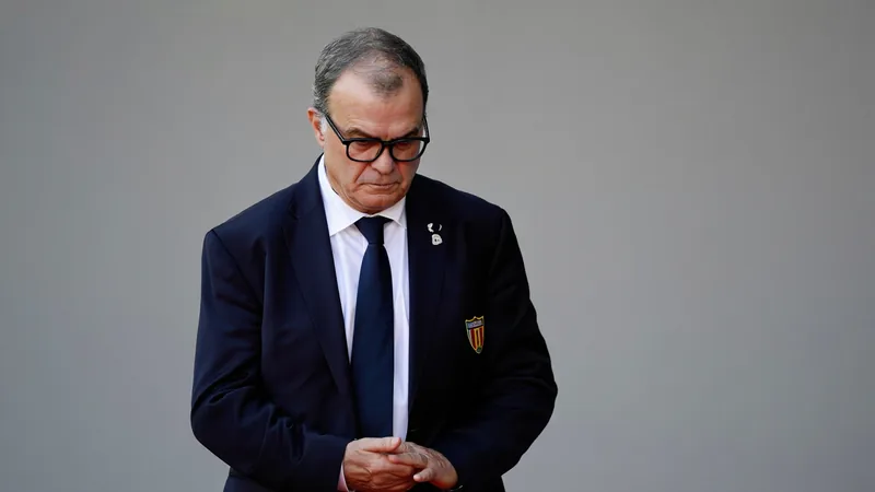 Marcelo Bielsa looking sternly downward during his official FIFA World Cup portrait photoshoot, wearing a dark suit and glasses.