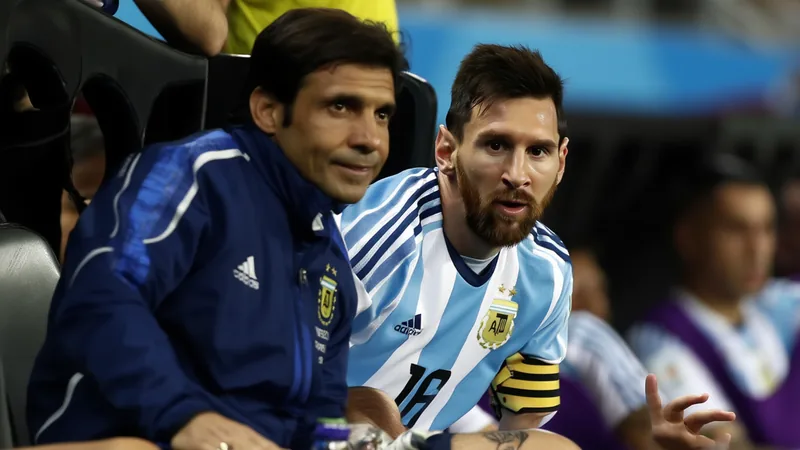 Pablo Aimar and Lionel Messi on the Argentina bench during a match, with Aimar in a coaching tracksuit and Messi in his playing kit.