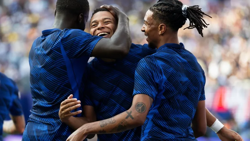 France national football team celebrating a goal, with Kylian Mbappe in the foreground and Didier Deschamps in the background.