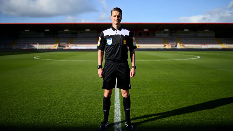 Rob Dieperink, a Dutch football referee in his officiating kit, standing on a soccer field with a stadium in the background.