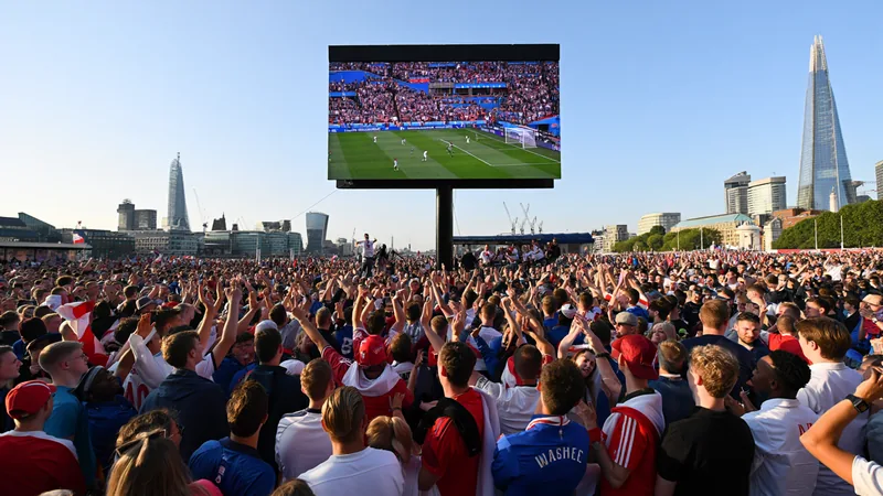 Fans watching England's World Cup match on a giant screen at an outdoor fan zone in London, with city skyline in background.