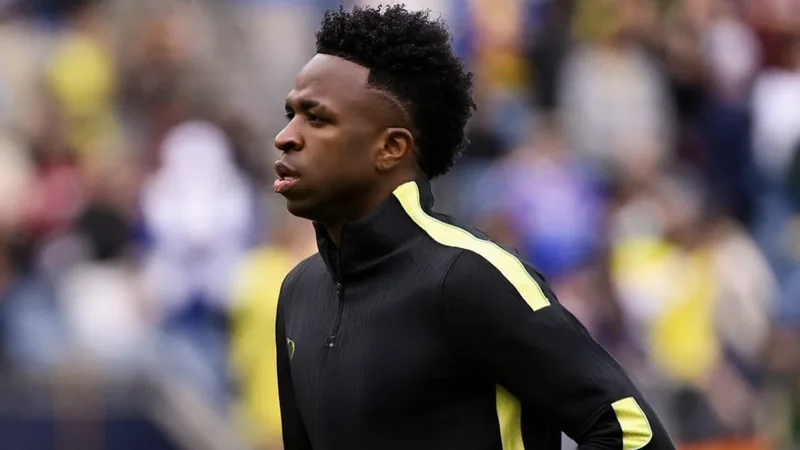 Vinícius Júnior im brasilianischen Nationaltrikot, jubelnd mit erhobenen Armen auf dem Spielfeld, im Hintergrund die brasilianische Flagge.