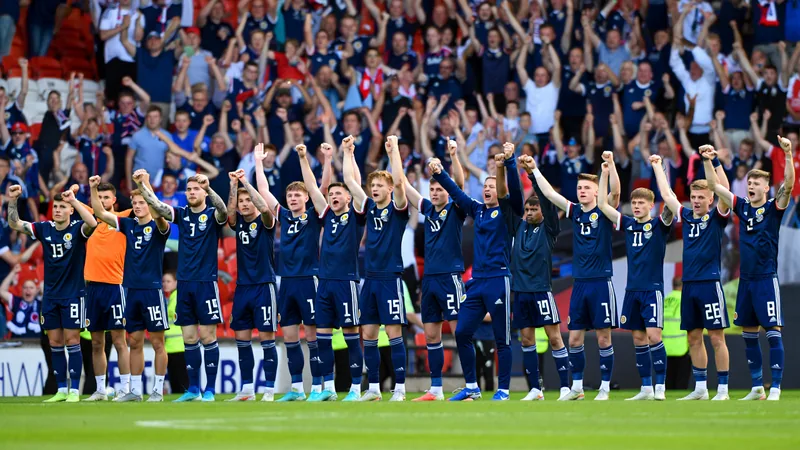 Was bedeutet es, für Schottland bei einer WM zu spielen? — Football Schottische Fußballspieler in dunkelblauen Trikots jubeln gemeinsam auf dem Rasen, im Hintergrund die schottische Flagge und ein Stadion.