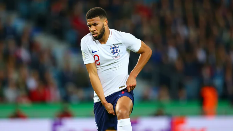 Guehi droht Ausfall gegen Norwegen — Football World Cup News Marc Guehi im englischen Trikot auf dem Rasen des Aztekenstadions, hält sich den Oberschenkel, Trainer Thomas Tuchel und Teamkollegen im Hintergrund.