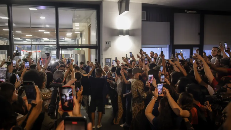 Santiago Castro lächelt am Flughafen Rom-Fiumicino, im Hintergrund das AS-Rom-Logo, in der Hand einen Schal des Vereins.