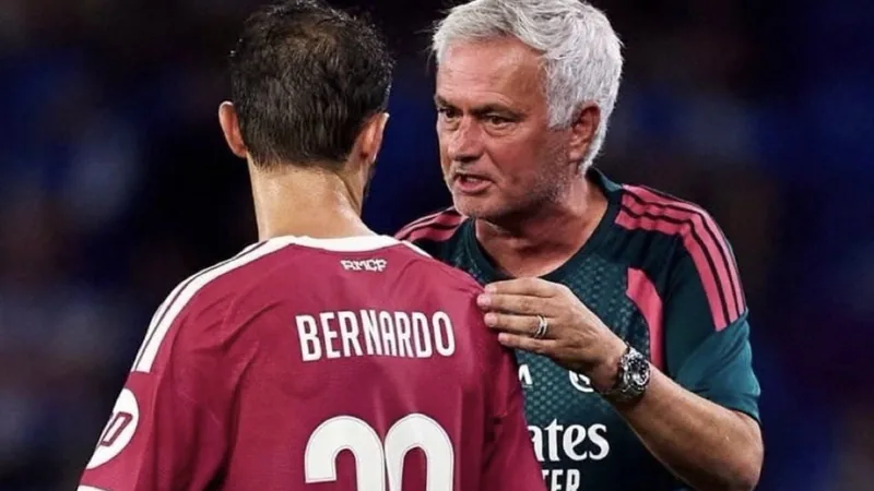 José Mourinho in einem Trainingsanzug, der auf einer Pressekonferenz lächelt, während im Hintergrund das Logo von Manchester City zu sehen ist.