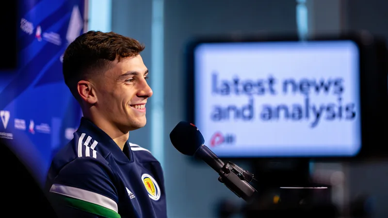 Ryan Christie con la camiseta de Escocia sonriendo en una entrevista, con el campo de fútbol de fondo y compañeros de equipo desenfocados.