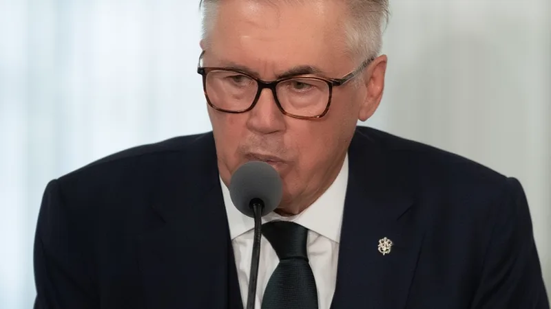 Carlo Ancelotti en la rueda de prensa del Real Madrid, con gesto serio, vistiendo traje y camisa blanca, micrófonos y periodistas al fondo.