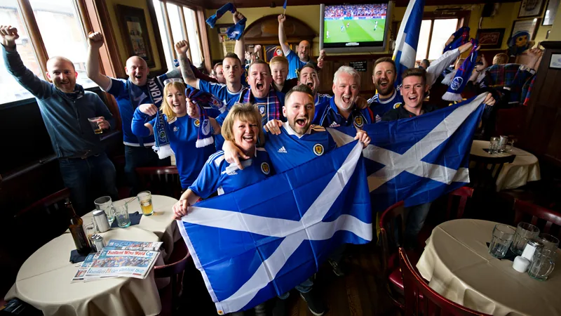 Aficionados escoceses celebrando en un pub de Corby, con banderas de Escocia y camisetas de la selección, frente a una pantalla gigante que muestra