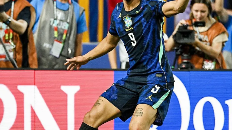 Matheus Cunha celebrando un gol con la camiseta de Brasil en un estadio lleno de aficionados, con el marcador de fondo mostrando el resultado del