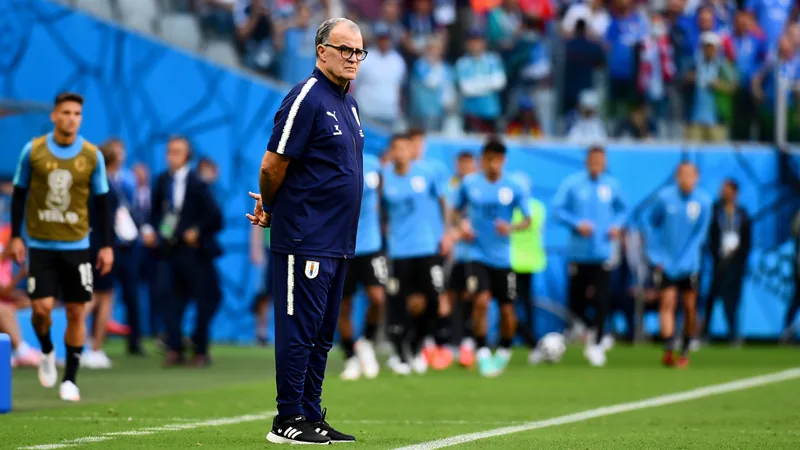 Marcelo Bielsa de pie en el área técnica durante un partido de Uruguay, con expresión seria y brazos cruzados, vistiendo traje oscuro.