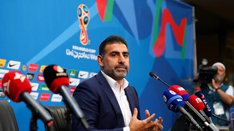 Amir Ghalenoei, entrenador de Irán, hablando en una conferencia de prensa con gesto serio, con el logo de la Copa del Mundo de fondo.