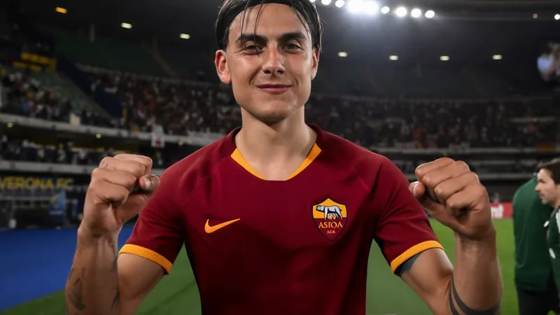 La Roma recibe la contrapropuesta de Dybala para renovar — Football Paulo Dybala con la camiseta de la Roma celebrando un gol en el Estadio Olímpico de Roma, con aficionados de fondo y banderas del club