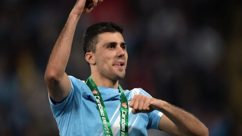 Manchester City ofrece nuevo contrato a Rodri, pero aún sin acuerdo Rodri, centrocampista del Manchester City, celebrando un gol con el puño en alto, con el estadio Etihad de fondo y aficionados vitoreando.