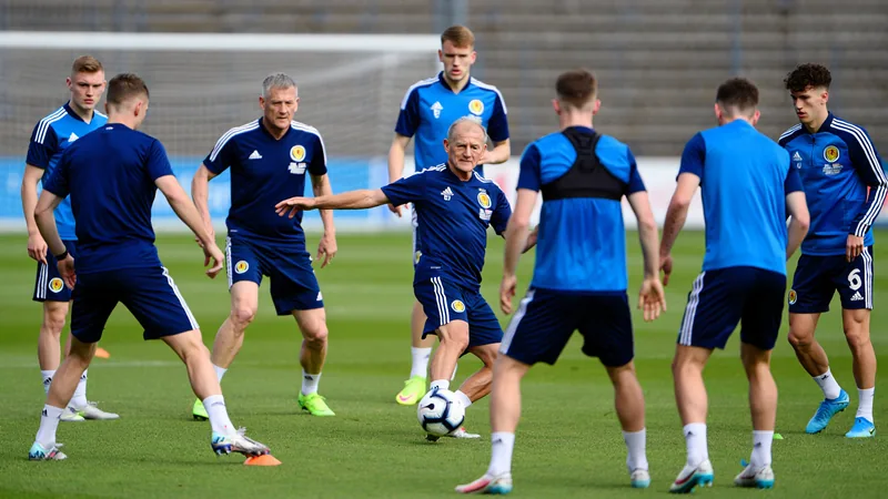 El complicado reto del nuevo seleccionador de Escocia — Football Un entrenador de fútbol con una carpeta en la mano observa a jugadores escoceses entrenando en un campo verde, con banderas de Escocia al fondo.