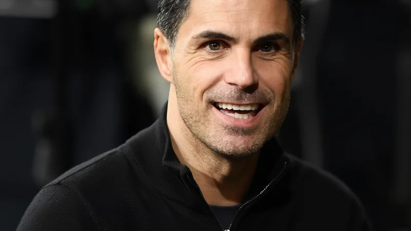 Arteta defiende a Lewis-Skelly ante especulaciones — Football World Mikel Arteta en la banda del Emirates Stadium durante un partido del Arsenal, con gesto serio y brazos cruzados, mientras los jugadores calientan al