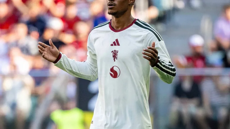 Marcus Rashford con la camiseta del Manchester United sonriendo en el campo de entrenamiento de Carrington, con el escudo del club al fondo.