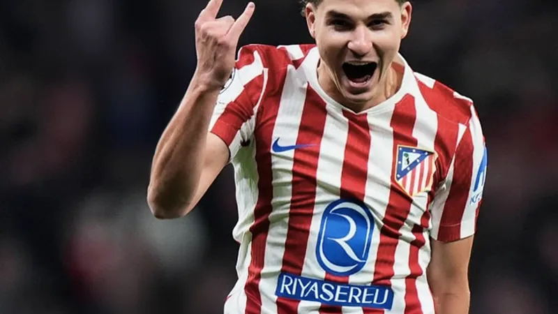 Julián Alvarez en tenue de l'Atlético Madrid célébrant un but, avec le logo du club en arrière-plan et des supporters en liesse.