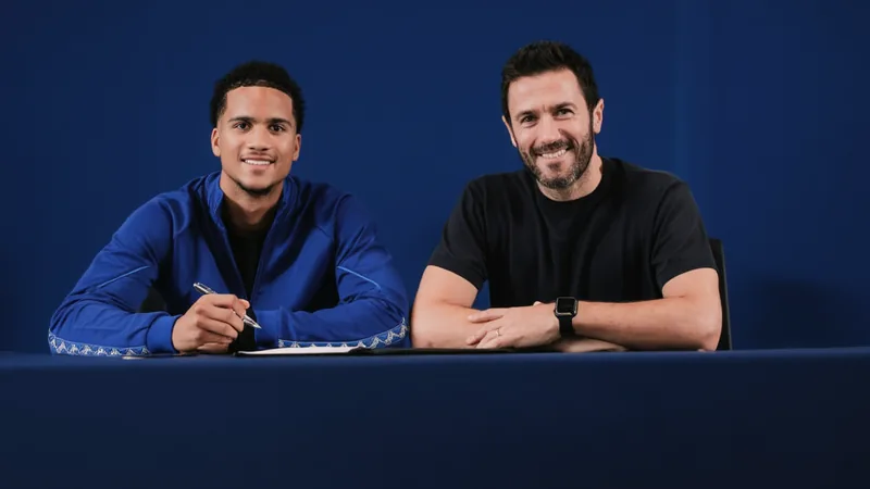 Divine Mukasa prolonge à Manchester City jusqu'en 2030 — Football Un jeune joueur de football en tenue de Manchester City, souriant, avec le logo du club en arrière-plan et un ballon à ses pieds.