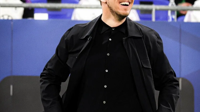Julian Nagelsmann en costume, debout sur un terrain de football, regardant pensivement l'horizon, avec des joueurs allemands en arrière-plan.