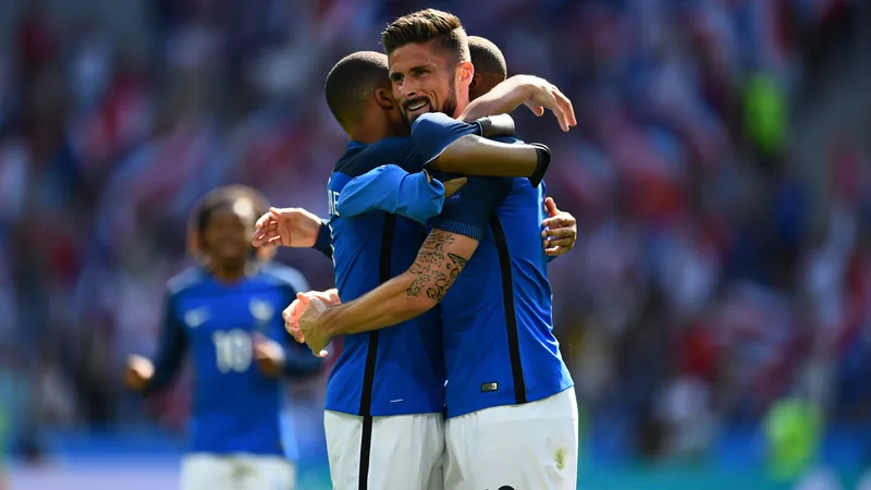 Olivier Giroud et Kylian Mbappé s'enlaçant sur un terrain de football après un but, avec des supporters en arrière-plan et un stade illuminé.