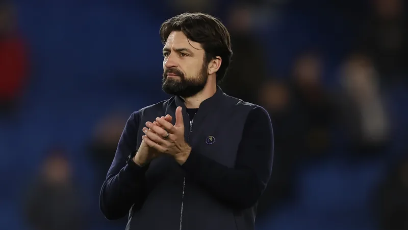 Russell Martin favori pour entraîner Leicester City — Football World Russell Martin, entraîneur de football, en costume, souriant sur un terrain d'entraînement avec un ballon de football à ses pieds