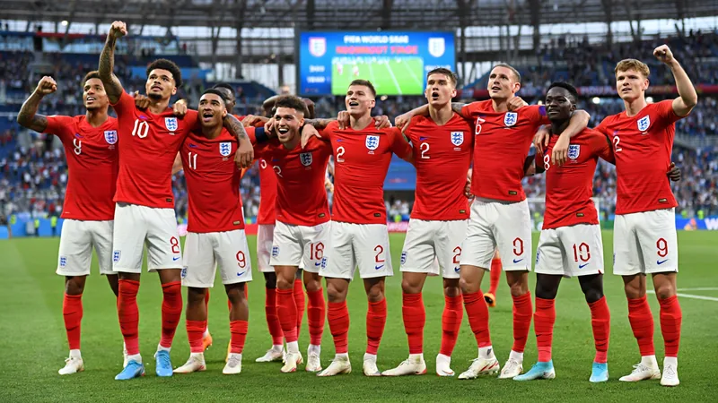 Joueurs de l'équipe d'Angleterre célébrant une victoire sur un terrain de football, avec le drapeau anglais agité par des supporters en arrière-plan.
