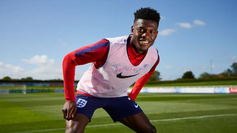 Ngumoha quitte la sélection anglaise après avoir marqué les esprits Rio Ngumoha en tenue d'entraînement de l'équipe d'Angleterre, souriant sur un terrain de football, avec le logo des Three Lions en arrière-plan.