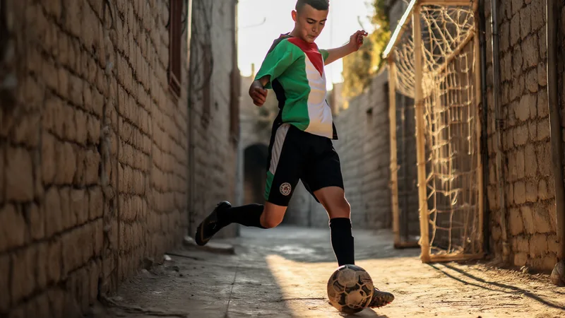 Football en Cisjordanie : une échappatoire pour les Palestiniens Jeunes Palestiniens jouant au football sur un terrain improvisé en Cisjordanie, avec des bâtiments et des collines en arrière-plan au coucher du