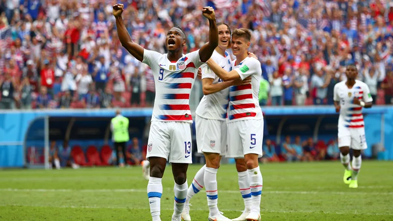Joueurs américains célébrant un but lors d'un match de Coupe du monde, avec le drapeau américain agité par les supporters en arrière-plan.