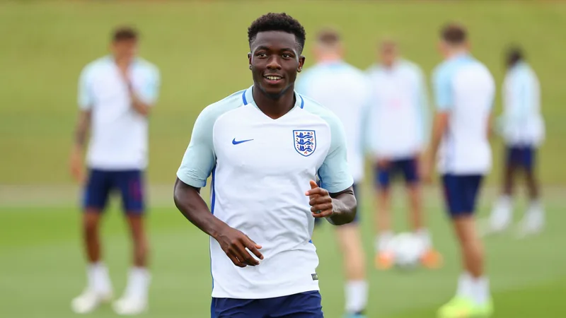 Bukayo Saka en tenue de l'Angleterre, souriant sur le terrain d'entraînement, avec le logo de la Coupe du monde en arrière-plan.