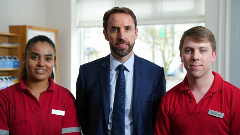 Sir Gareth Southgate souriant avec deux employées de bureau de poste derrière le comptoir, avec des timbres et une caisse enregistreuse visibles.