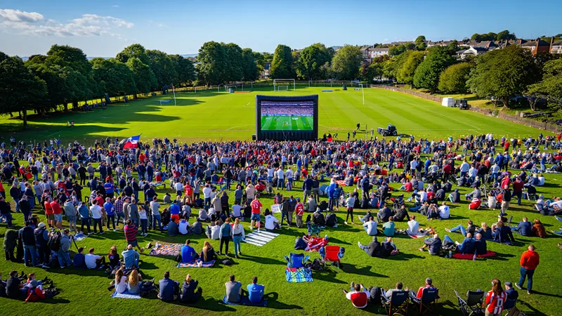 Vue d'Abbey Park à Torquay avec un grand écran installé sur la pelouse, des supporters anglais en liesse brandissant des drapeaux, et la mer en
