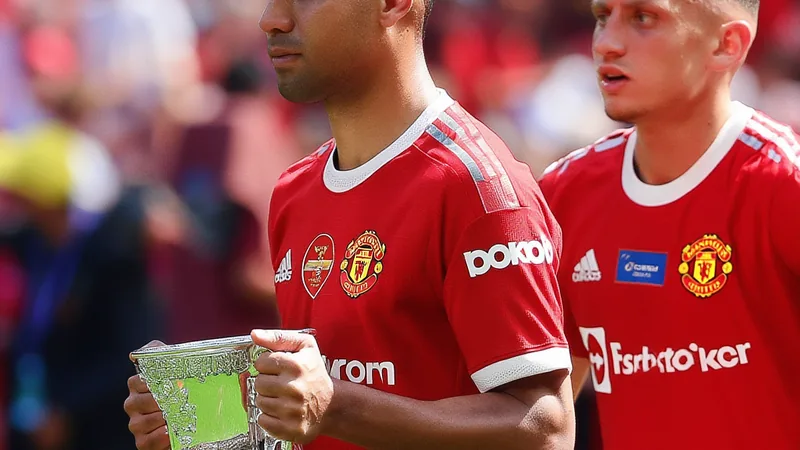 Tyrell Malacia et Casemiro quittent Manchester United — Football Tyrell Malacia et Casemiro en tenue de Manchester United quittant le stade Old Trafford, avec le logo du club en arrière-plan.