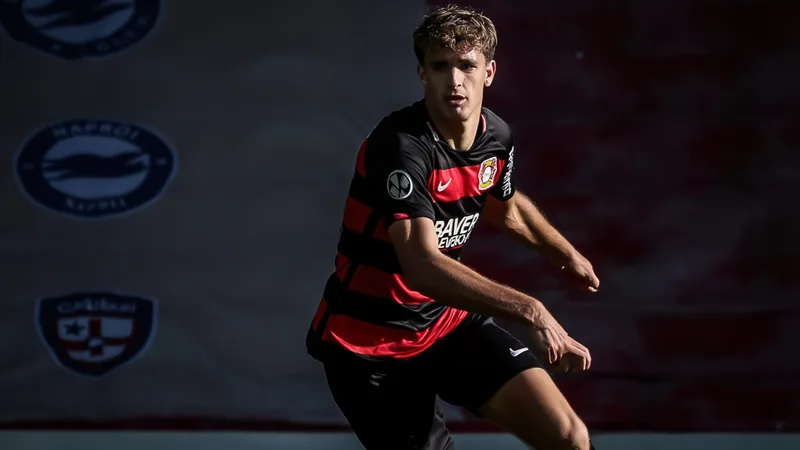 Un jeune défenseur central en action lors d'un match de Bundesliga, portant le maillot du Bayer Leverkusen, avec des recruteurs en tribune.