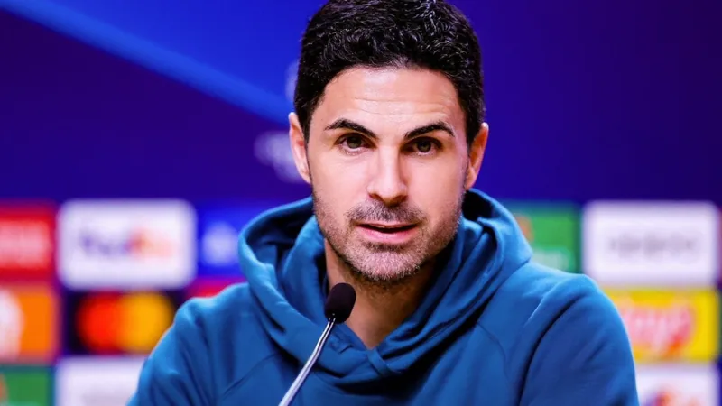 Mikel Arteta, entraîneur d'Arsenal, en costume, regardant un match depuis la touche, avec des joueurs en arrière-plan et le stade de l'Emirates