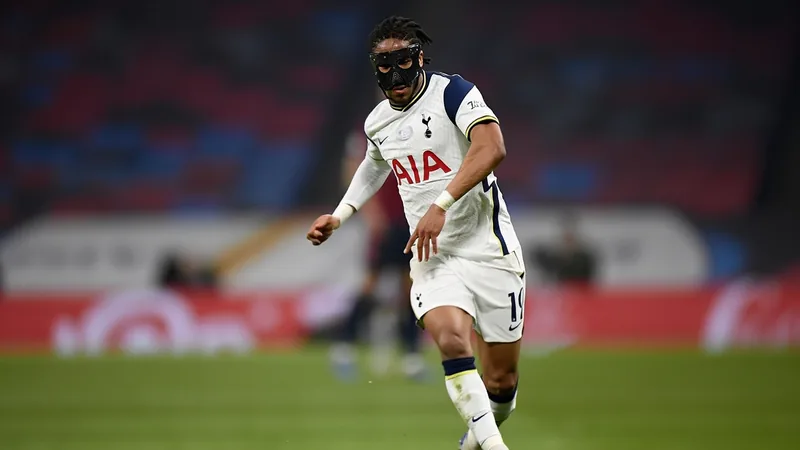 Jogador Djed Spence do Tottenham com máscara protetora facial em campo, durante partida de futebol, com torcida ao fundo.