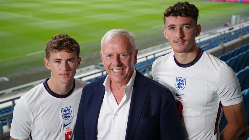 Nigel James, pai orgulhoso, abraçando seus dois filhos vestindo uniformes da seleção inglesa de futebol, em um campo de grama verde.