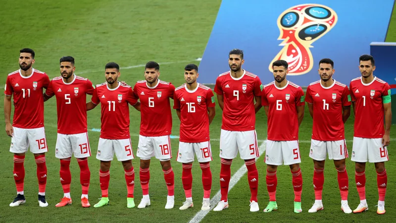 Jogadores da seleção iraniana de futebol em campo, com bandeiras do Irã ao fundo, durante partida da Copa do Mundo de 2026.