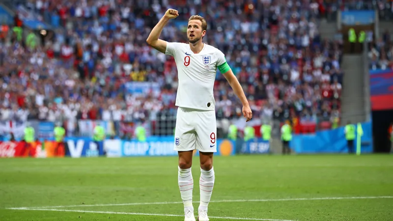 Harry Kane comemora um gol pela Inglaterra em uma partida da Copa do Mundo, vestindo a camisa branca, com o estádio ao fundo e torcedores vibrando.