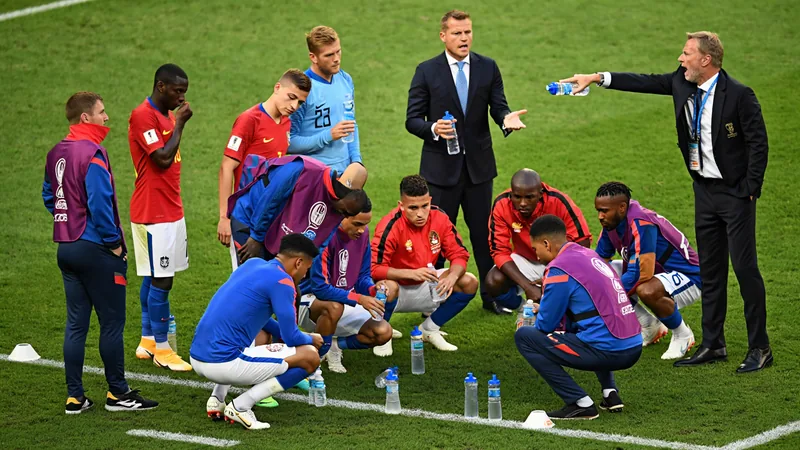 Pausas para hidratação no Mundial: quem ganha e quem perde Jogadores de futebol bebendo água durante uma pausa para hidratação em um estádio lotado, com o árbitro ao centro e placar ao fundo.