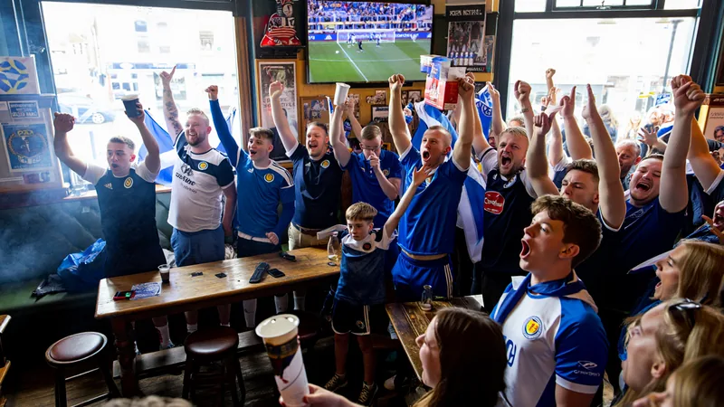 Torcida da 'Pequena Escócia' celebra vitória histórica — Football Torcedores escoceses vestidos com kilts e bandeiras da Escócia comemorando em uma rua de Corby, Inglaterra, com faixas e fogos de artifício ao fundo.