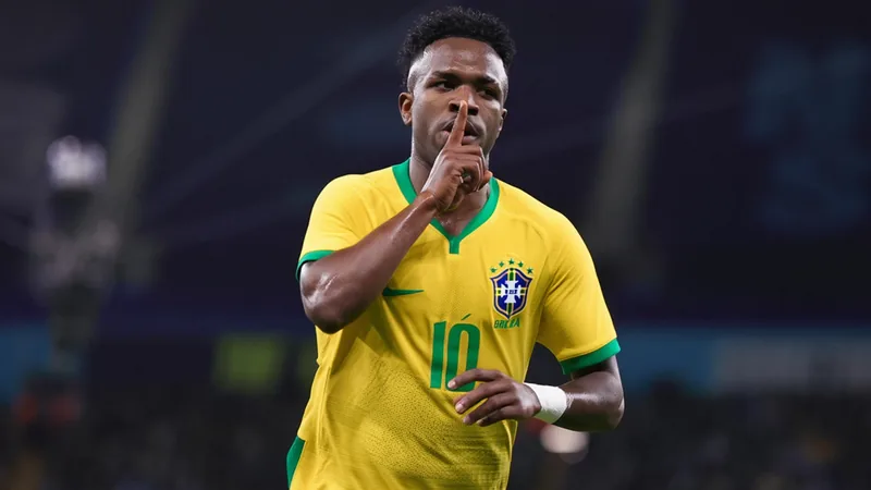Vinicius Junior com a camisa da seleção brasileira durante treino, sorrindo e olhando para a câmera, com o campo de futebol ao fundo.