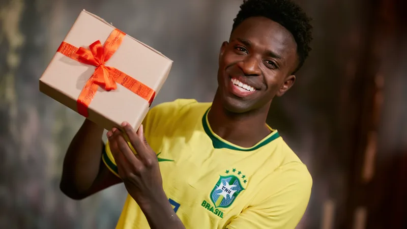 Vinícius Júnior com a camisa do Real Madrid, sorrindo durante entrevista coletiva, ao lado de uma imagem de Carlo Ancelotti no fundo