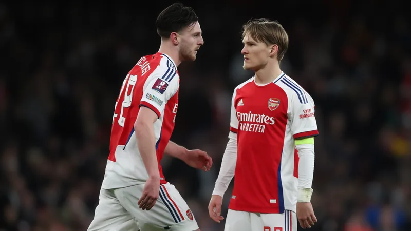 Declan Rice e Martin Odegaard, ambos com camisolas do Arsenal, frente a frente no centro do campo durante um jogo de futebol, com estádio lotado ao