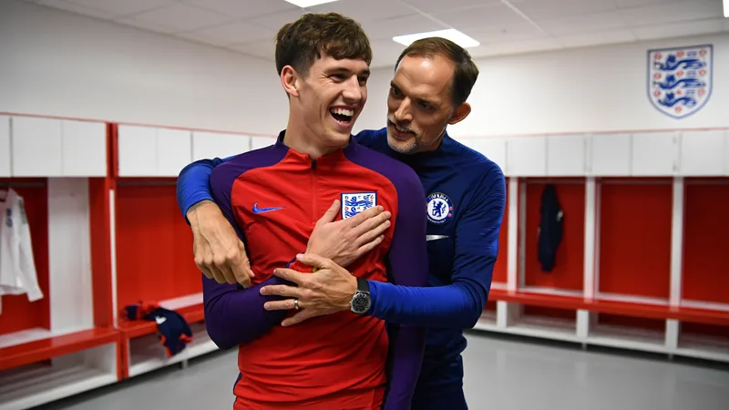 John Stones sobre pegadinha viral: 'Era boa vibração ali' — Football John Stones sorrindo enquanto segura o ombro em uma pegadinha no vestiário da Inglaterra, com Thomas Tuchel ao fundo parecendo preocupado.