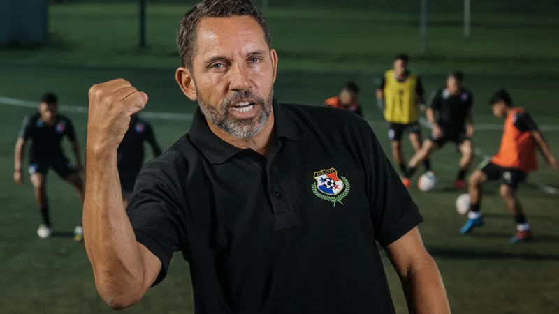 Mike Stump: Mundial Sub-20 seria enorme para o Panamá — Football Mike Stump, técnico do Panamá Sub-20, sorrindo durante um treino da seleção, com jogadores ao fundo em campo de futebol.