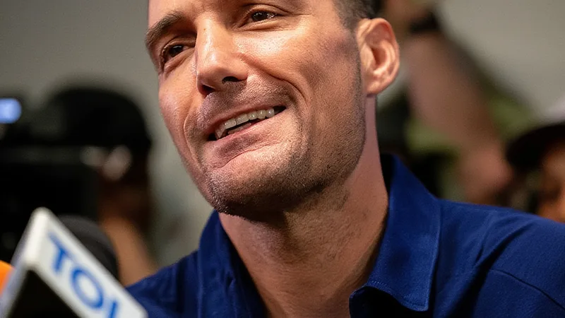 Lionel Scaloni sorrindo durante entrevista coletiva, com a camisa da seleção argentina, ao fundo bandeiras e logo da AFA.