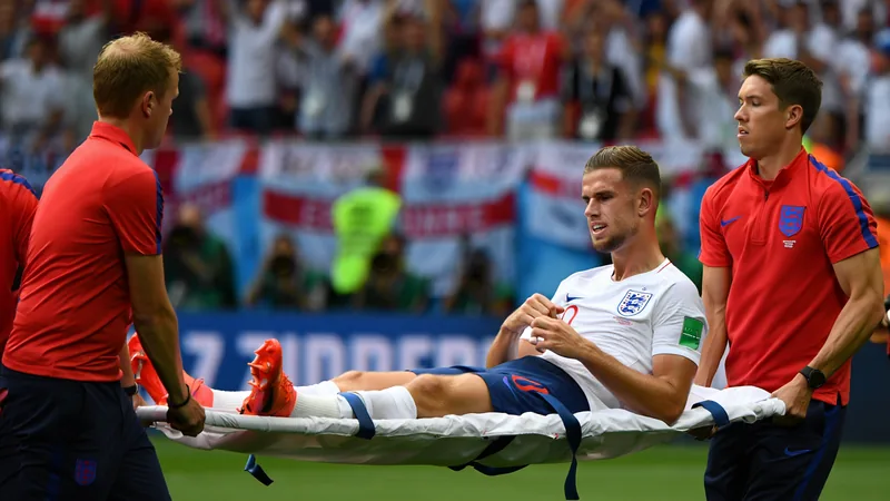 Jogadores de futebol ingleses celebrando em campo com painéis publicitários ao fundo, enquanto Jordan Henderson é levado de maca após cair ao tentar