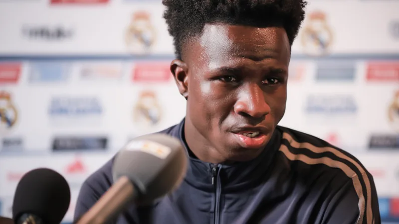 Vinicius Jr revela que Ancelotti decidiu dar pênalti para Bruno Vinicius Jr, do Real Madrid, falando em entrevista coletiva, com o logotipo da Champions League ao fundo e o estádio Santiago Bernabéu visível.